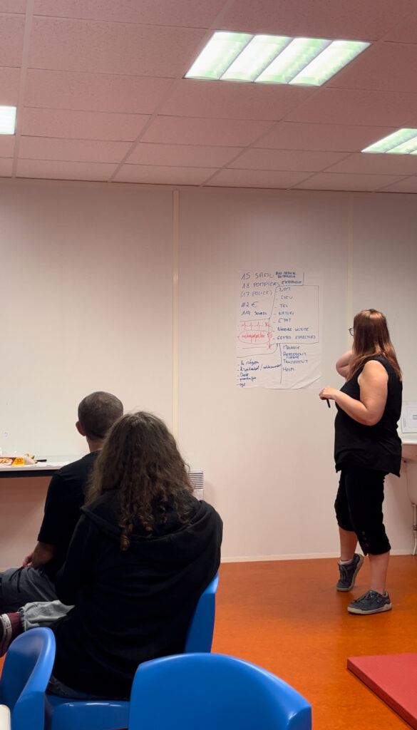 Sur un mur blanc une affiche avec une dame de c^té de dos en train de la lire, sur la gauche de l'image deux personnes assises tournées vers cette dame et la fiche en train d'écouter